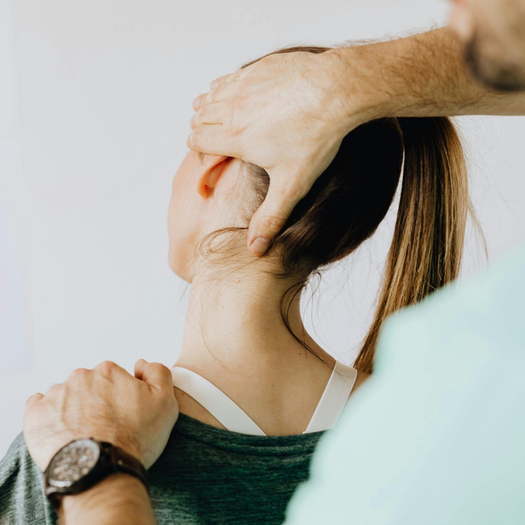 Back view of crop faceless bearded orthopedist in wristwatch checking up neck of anonymous lady while standing in front of wall with paper drawings representing body anatomy