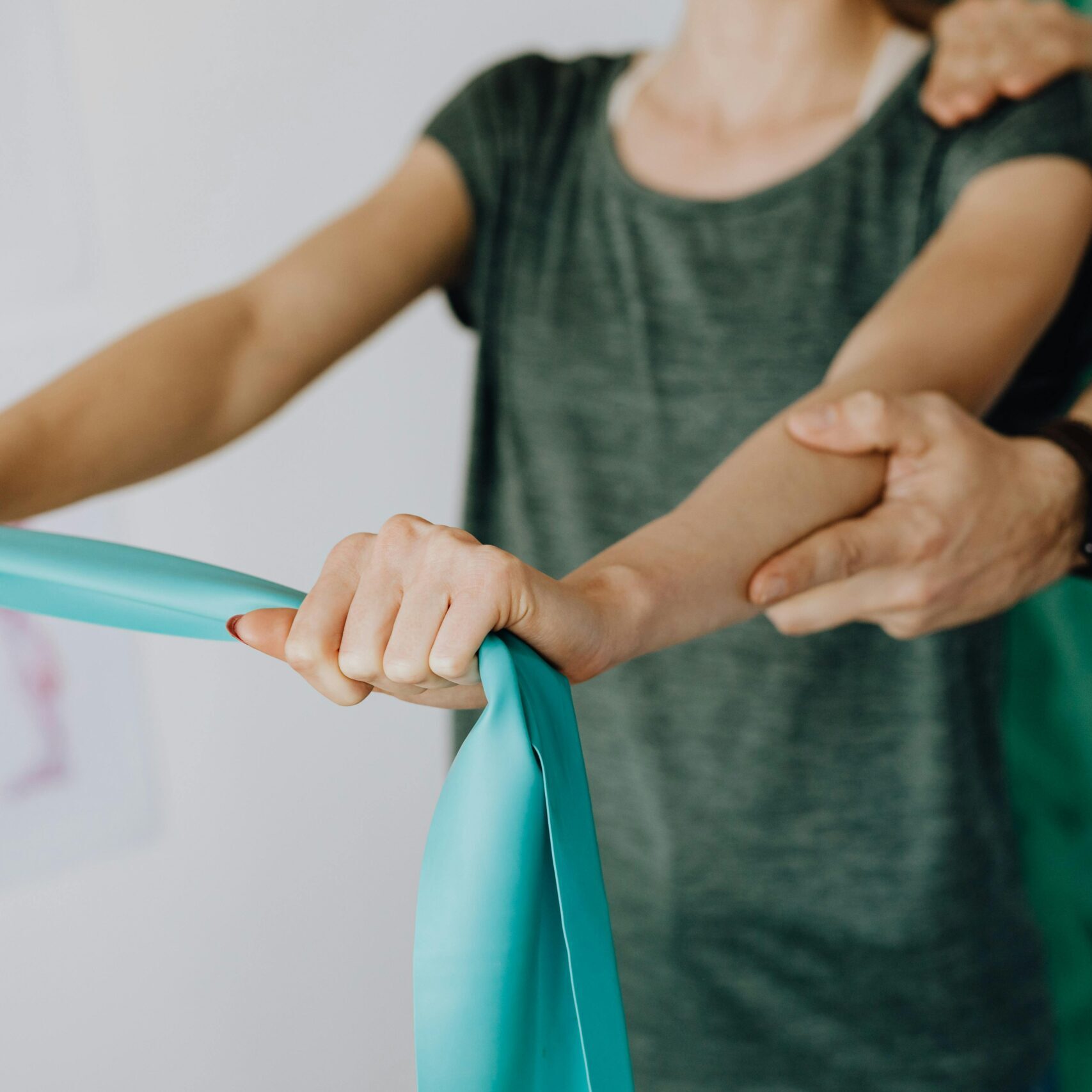 Crop unrecognizable orthopedist in uniform and wristwatch supporting shoulder and reached arm of faceless female patient with resistance band in doctor office