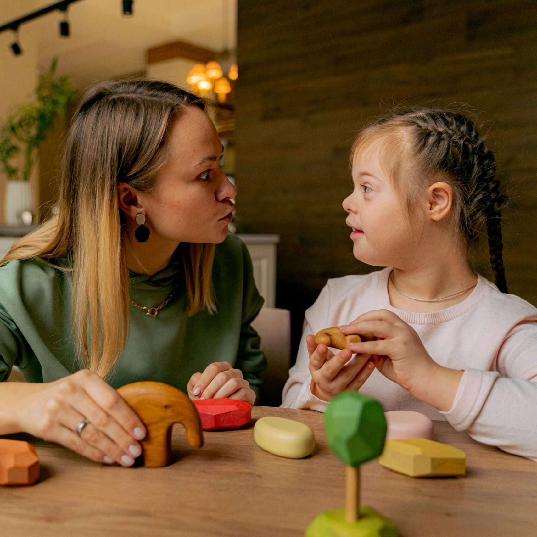 An adult woman and a child with Down syndrome engaging in playful interaction with wooden toys indoors.