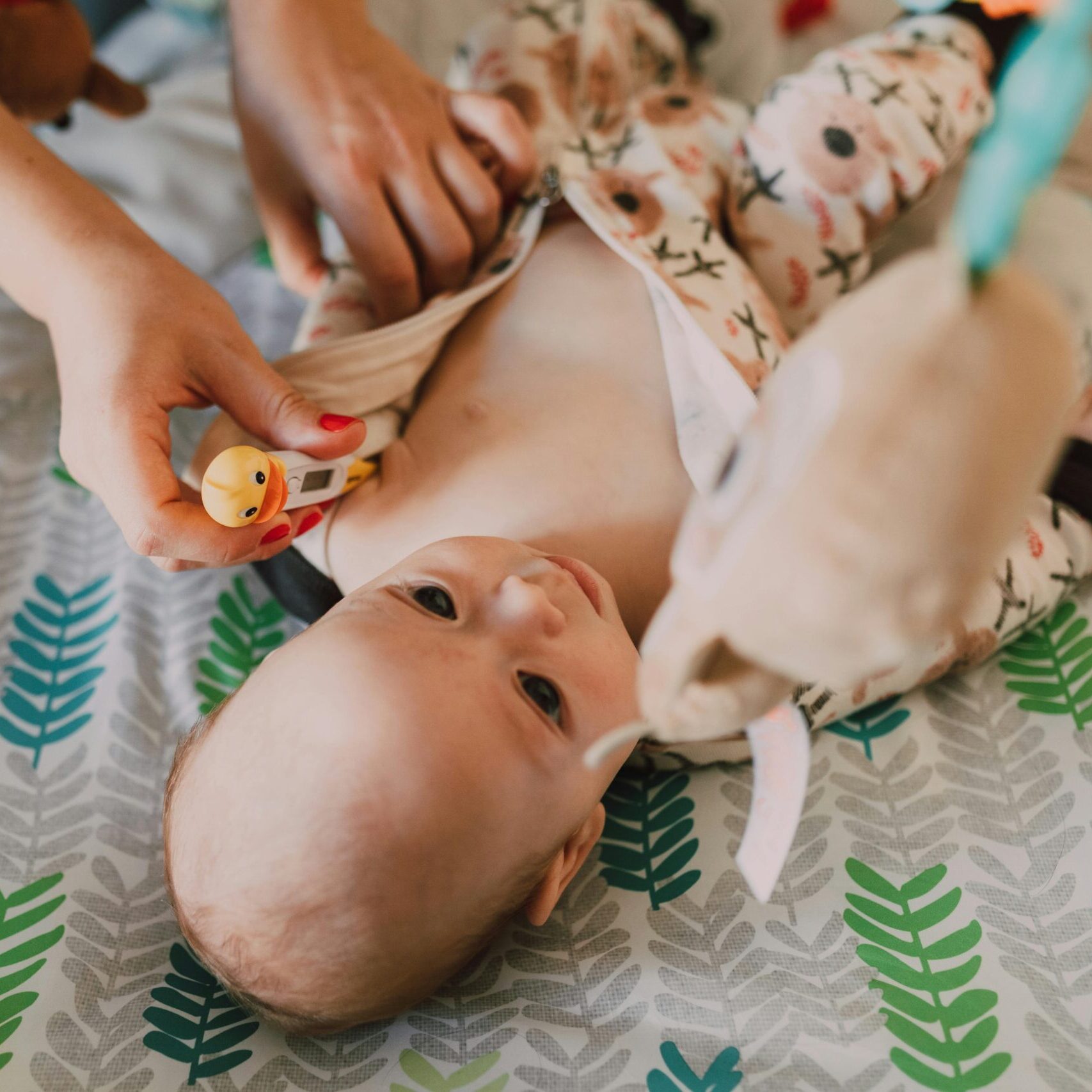 Parent using a thermometer for temperature check on a baby lying on a patterned bedspread.