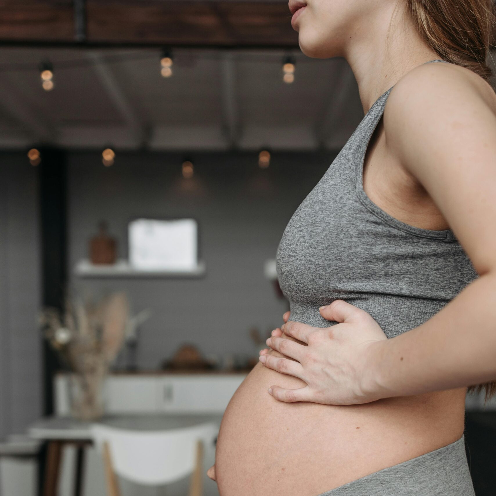 Side view of a pregnant woman wearing grey activewear, standing indoors in a cozy setting.