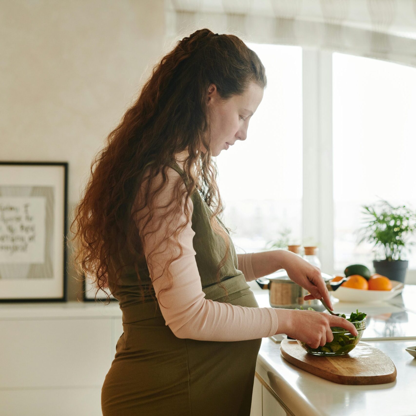 Pregnant woman with curly hair prepares a meal on kitchen counter, surrounded by fresh ingredients.