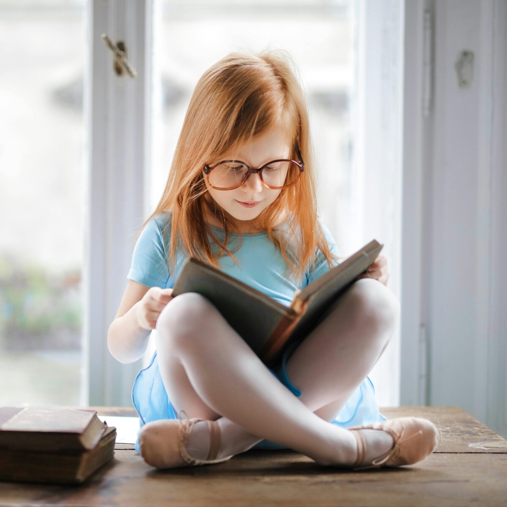 Adorable young girl wearing glasses, reading a book indoors by a window. Captures innocence and focus.