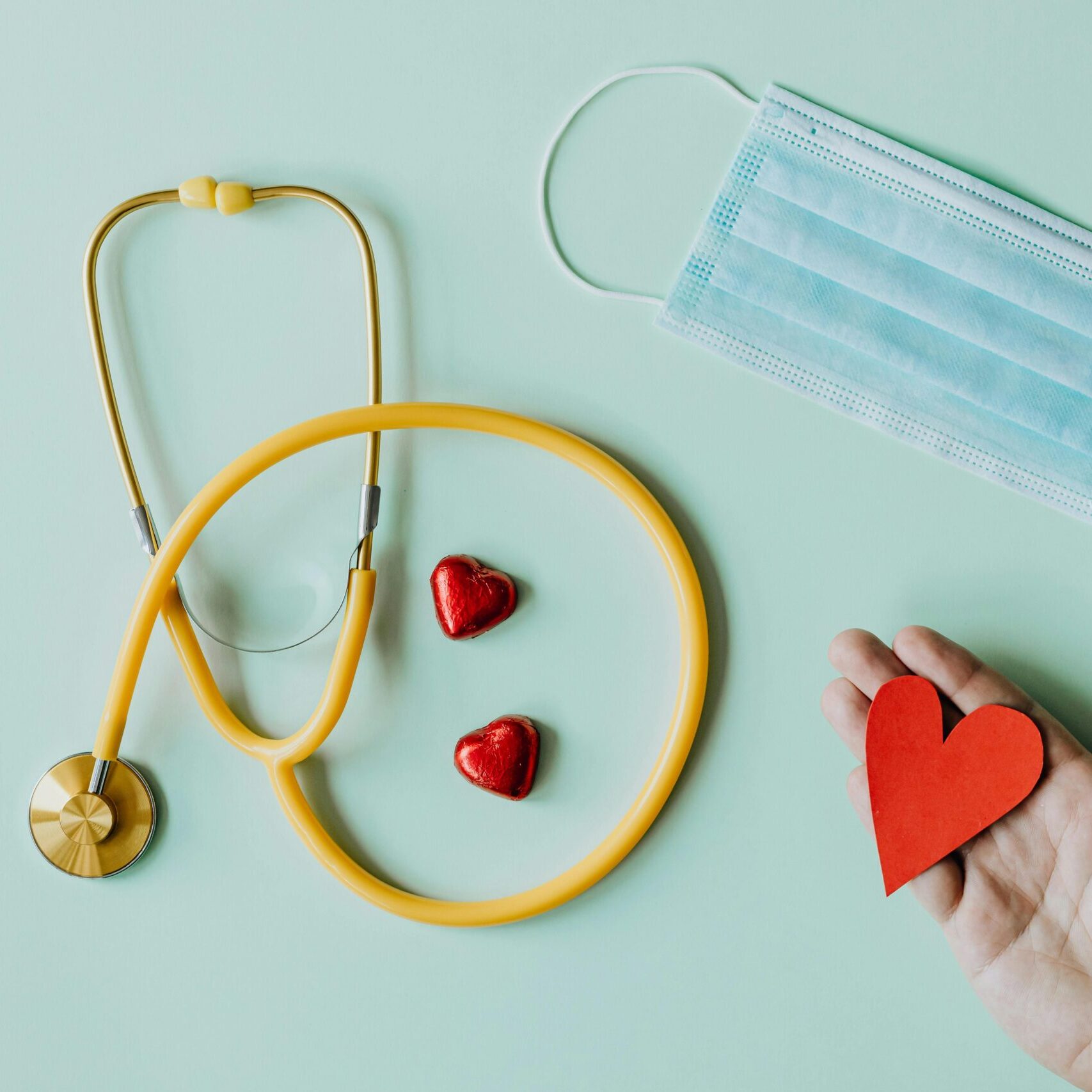 Top view of crop anonymous person hand with red paper heart on table with stethoscope and medical mask for coronavirus prevention