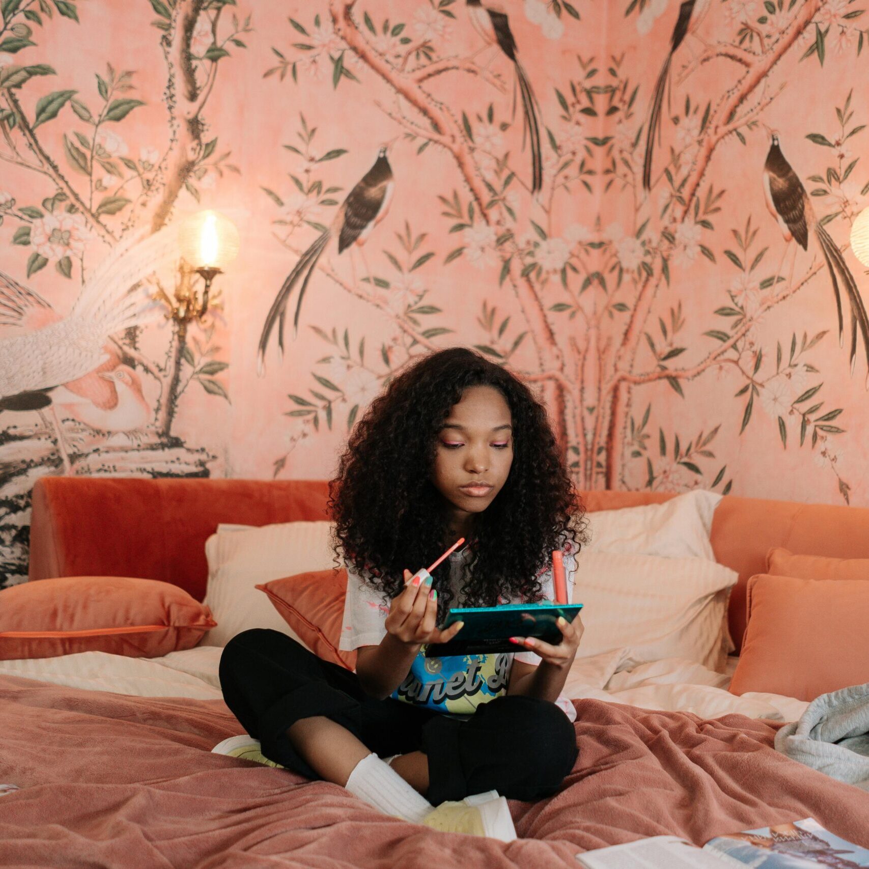 Young girl with curly hair applies makeup while sitting on a bed in a chic, artful bedroom. Cozy and fashionable setting.