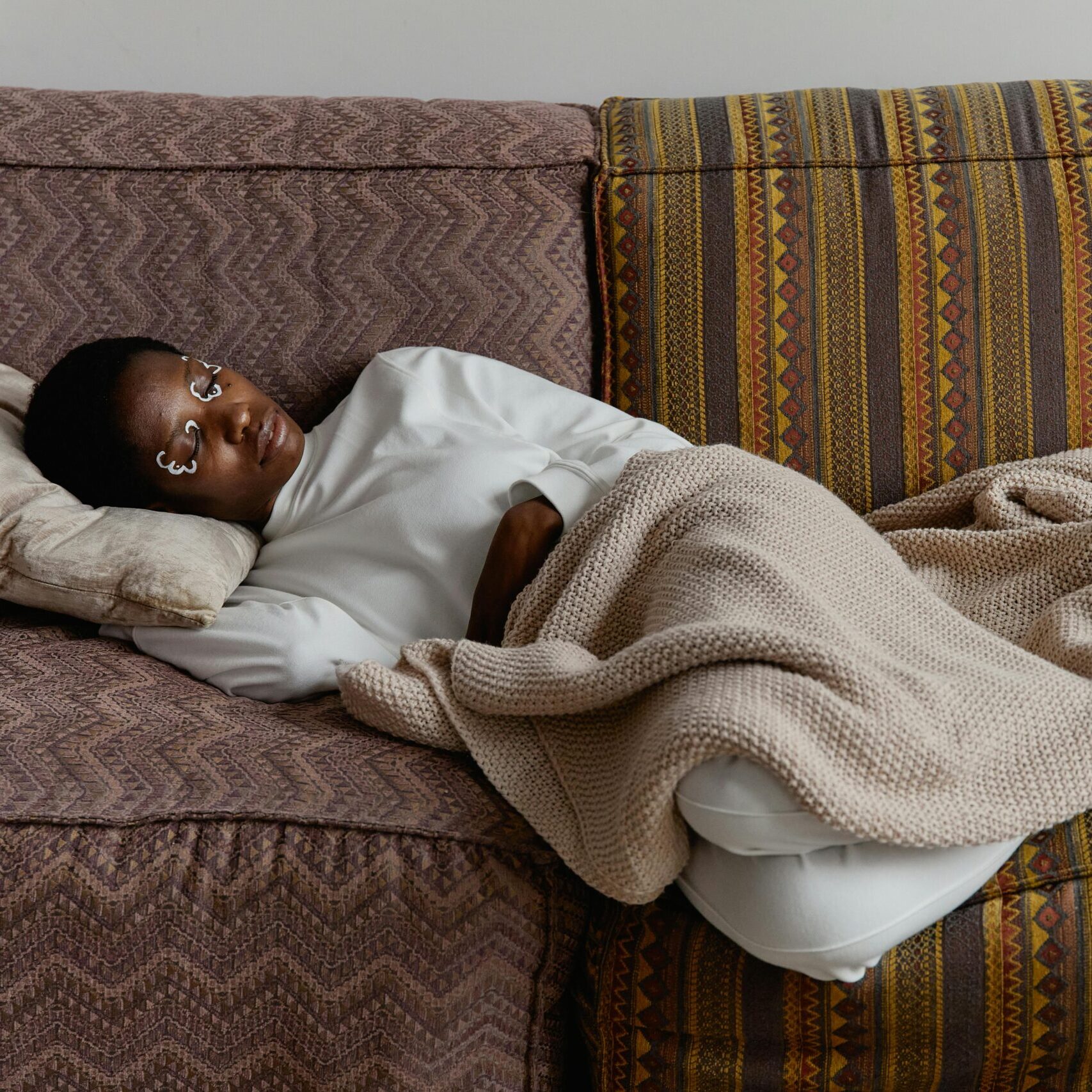 Fashionable young woman with face paint relaxing indoors on a cozy couch with blankets.